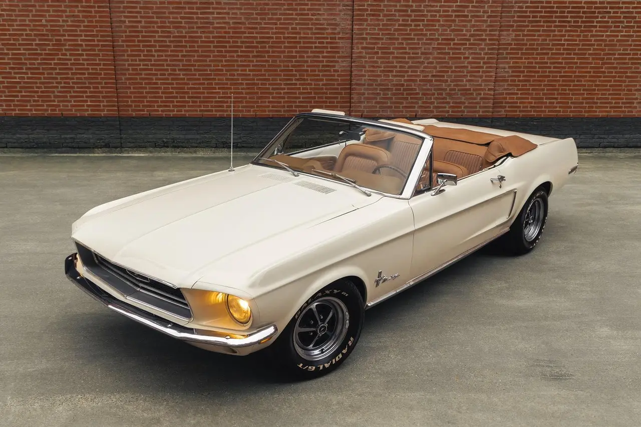 Crèmekleurige Ford Mustang-cabriolet uit 1968 met open kap en cognackleurig leren interieur, voor een bakstenen muur