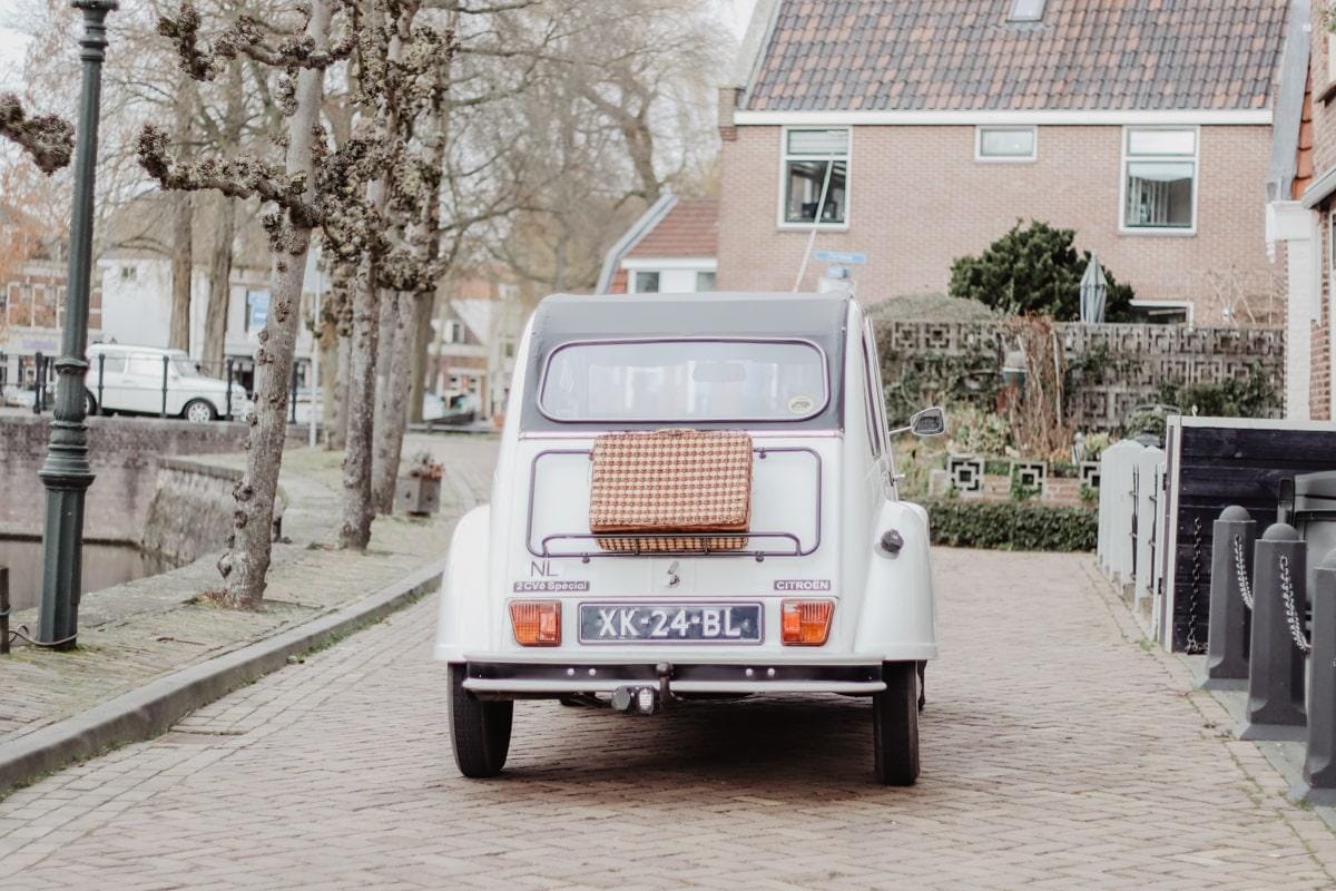 White Citroën 2CV seen from behind with a wicker suitcase on the luggage rack, on a cobbled street beside a canal