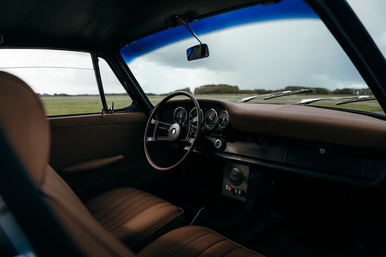 Interior of a classic Porsche with a slim steering wheel, wooden dashboard and cognac-coloured leather seats, with a grass field seen through the window