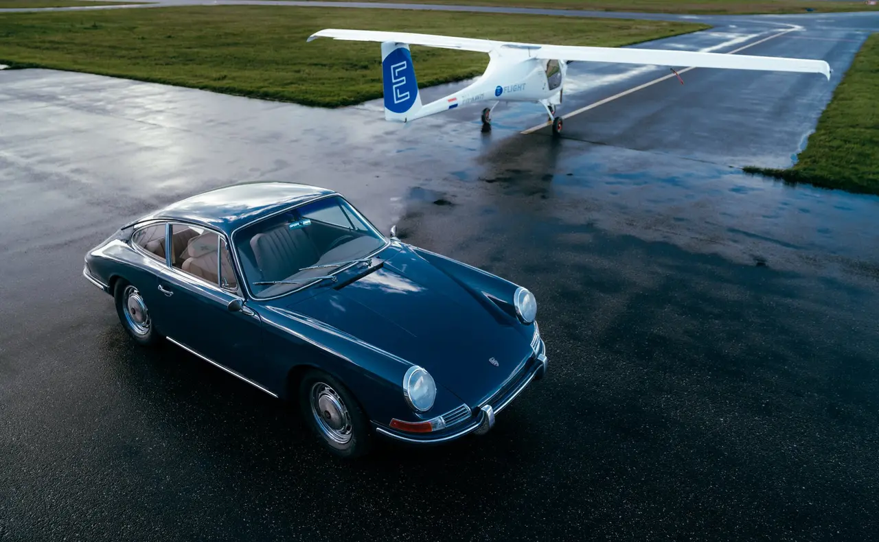 Bovenaanzicht van een donkerblauwe klassieke Porsche op een natte startbaan, met daarachter een wit elektrisch sportvliegtuigje op het gras