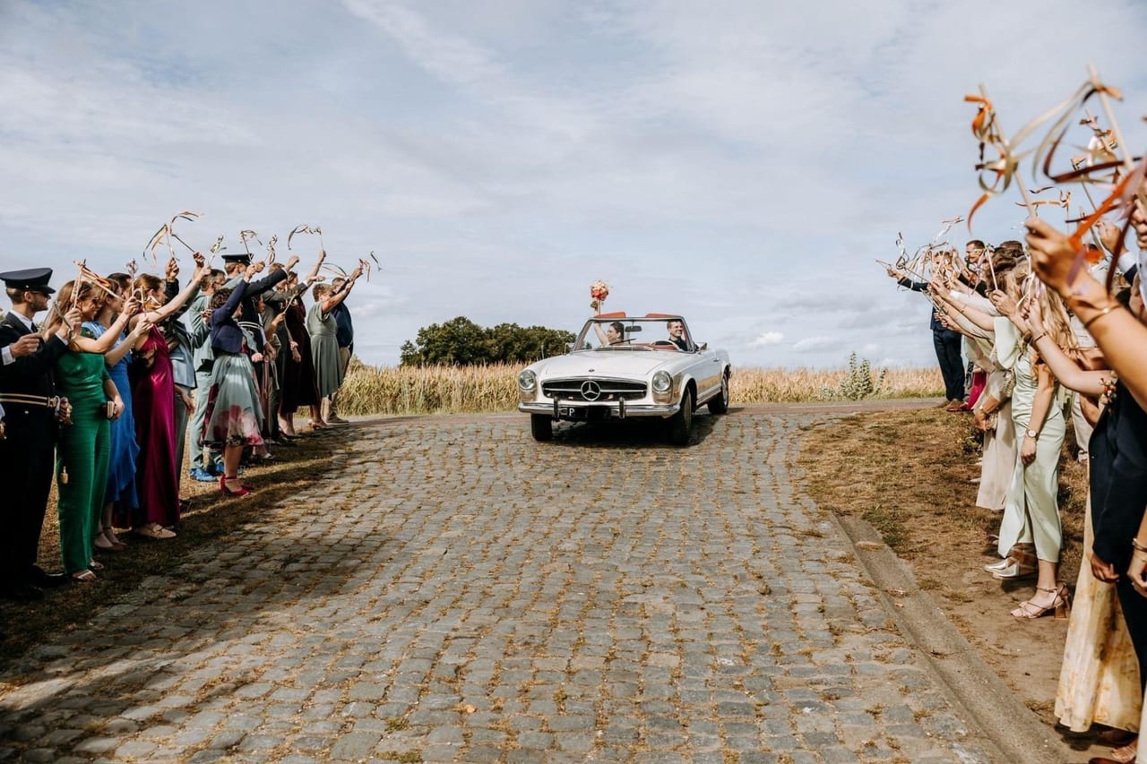 White Mercedes-Benz Pagode convertible with the bride and groom driving along a cobbled road beside a cornfield, between two rows of guests waving ribbons
