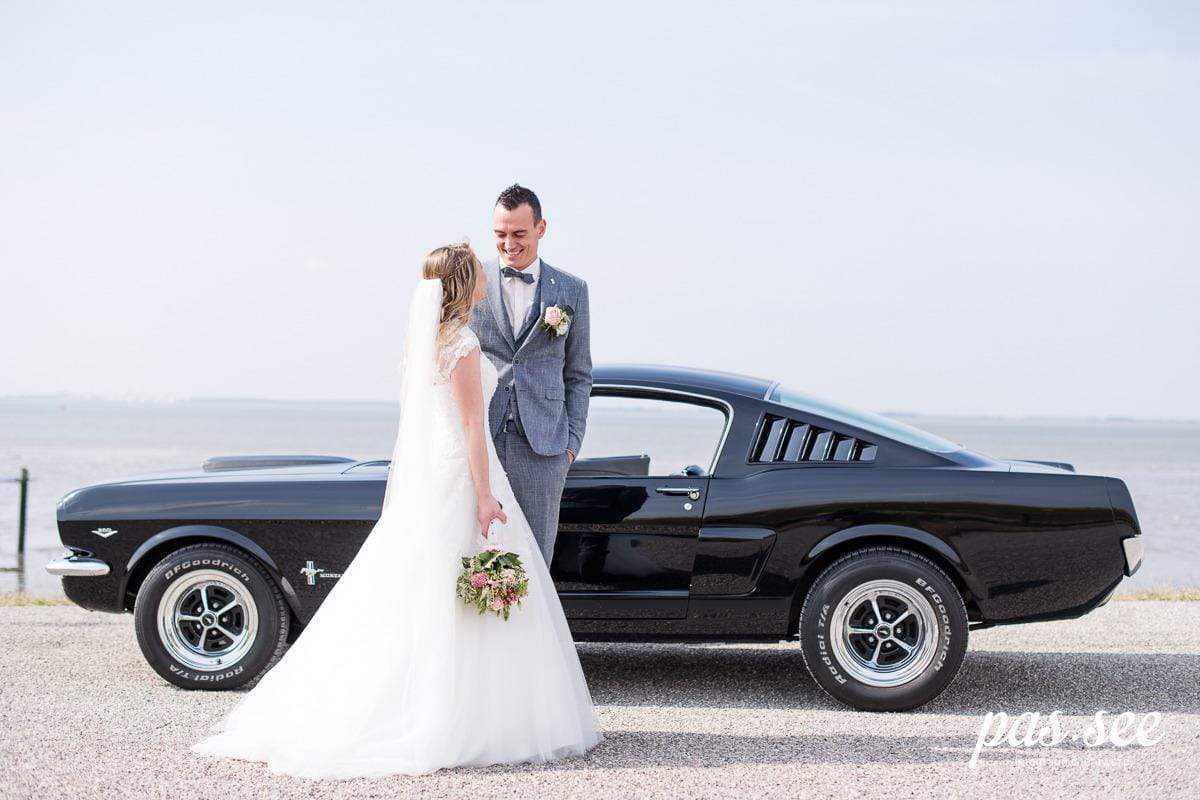 Bride in a long white dress with a veil and groom in a grey suit next to a low black classic Ford Mustang fastback, with water in the background