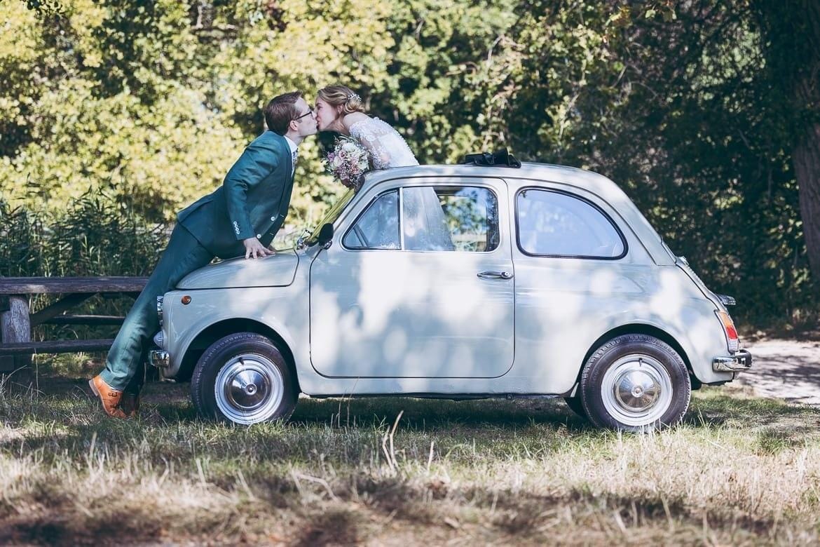 Couple kissing over the bonnet of a light grey classic Fiat 500 with the roof rolled back, on a grassy field in the sun