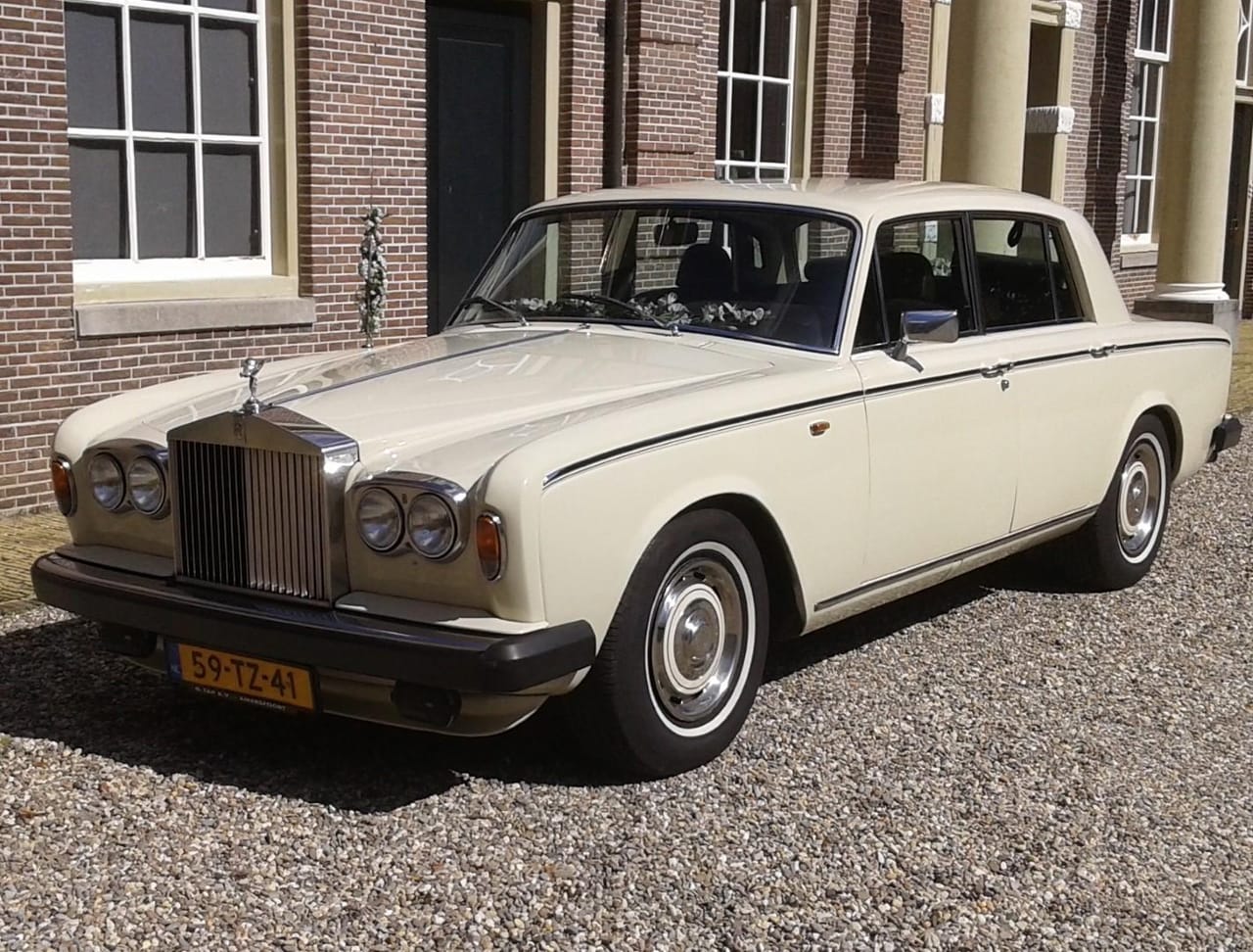 Crèmewitte Rolls-Royce Silver Shadow II op grind voor een bakstenen gebouw met hoge ramen en zuilen