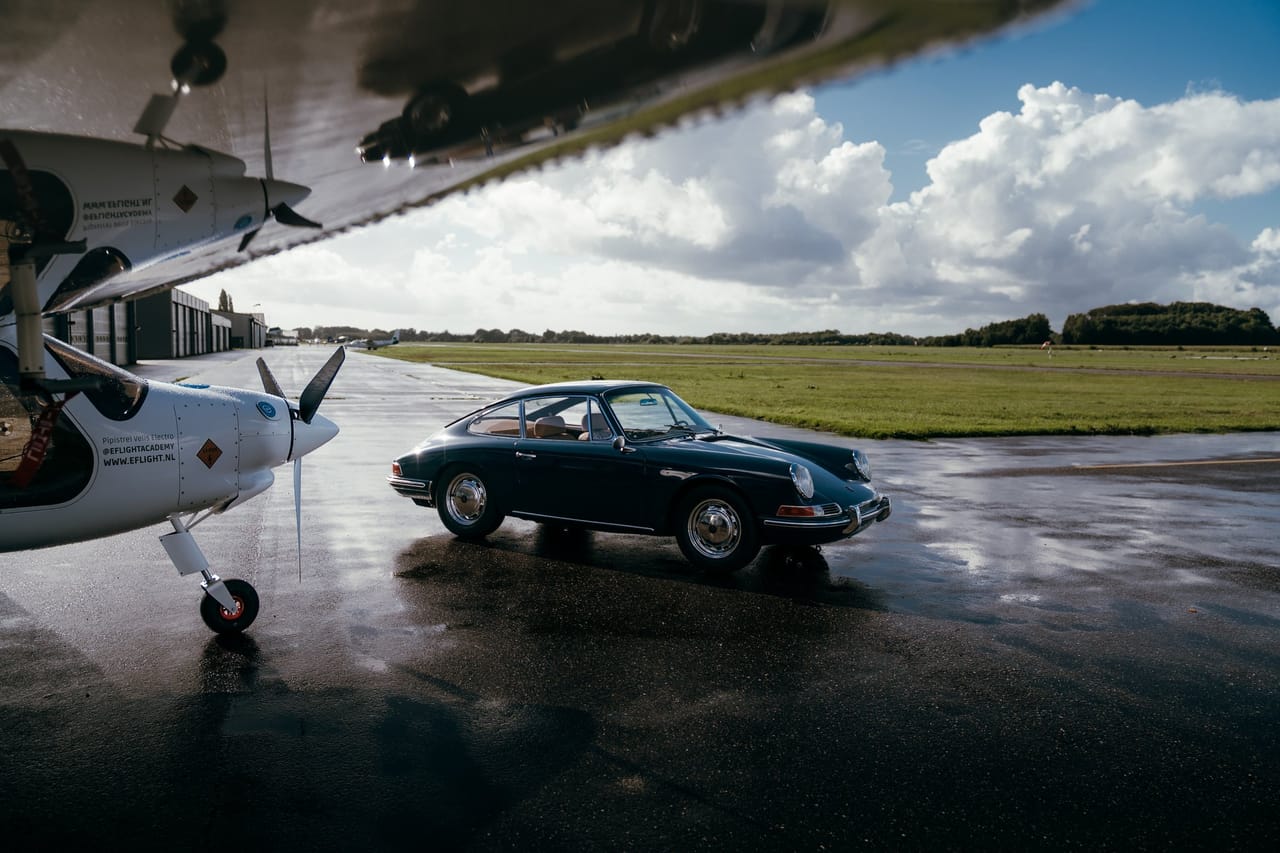 Dark blue classic Porsche on a wet apron next to a small propeller plane, seen from under the wing