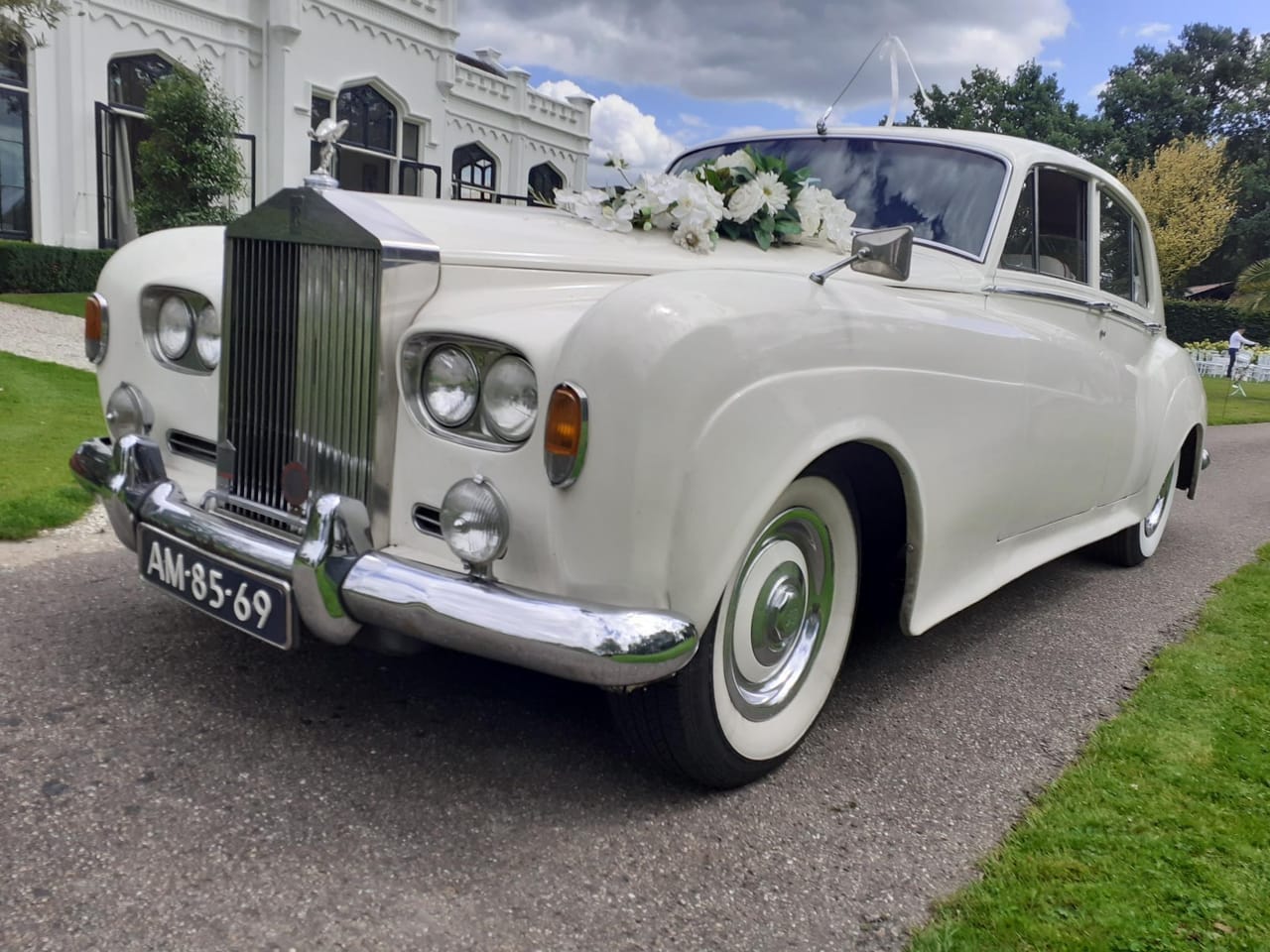 Witte Rolls-Royce Silver Cloud III met een bloemstuk van witte bloemen op de motorkap en een vrij kenteken voor, op een oprijlaan bij een wit landhuis