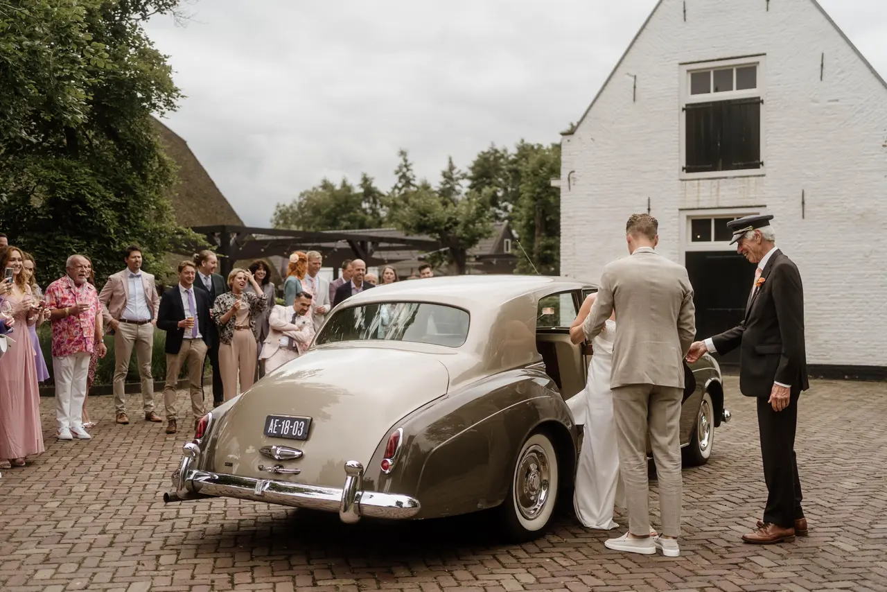 Chauffeur met pet houdt de deur open van een tweekleurige Rolls-Royce Silver Cloud terwijl het bruidspaar instapt, gasten kijken toe op een klinkerplein bij een witte boerderij