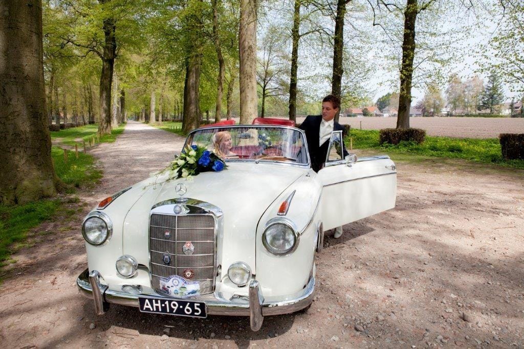 White Mercedes-Benz convertible with blue and white flowers on the bonnet, the bride sitting behind the windscreen and the groom getting in, on a sandy track between tall trees