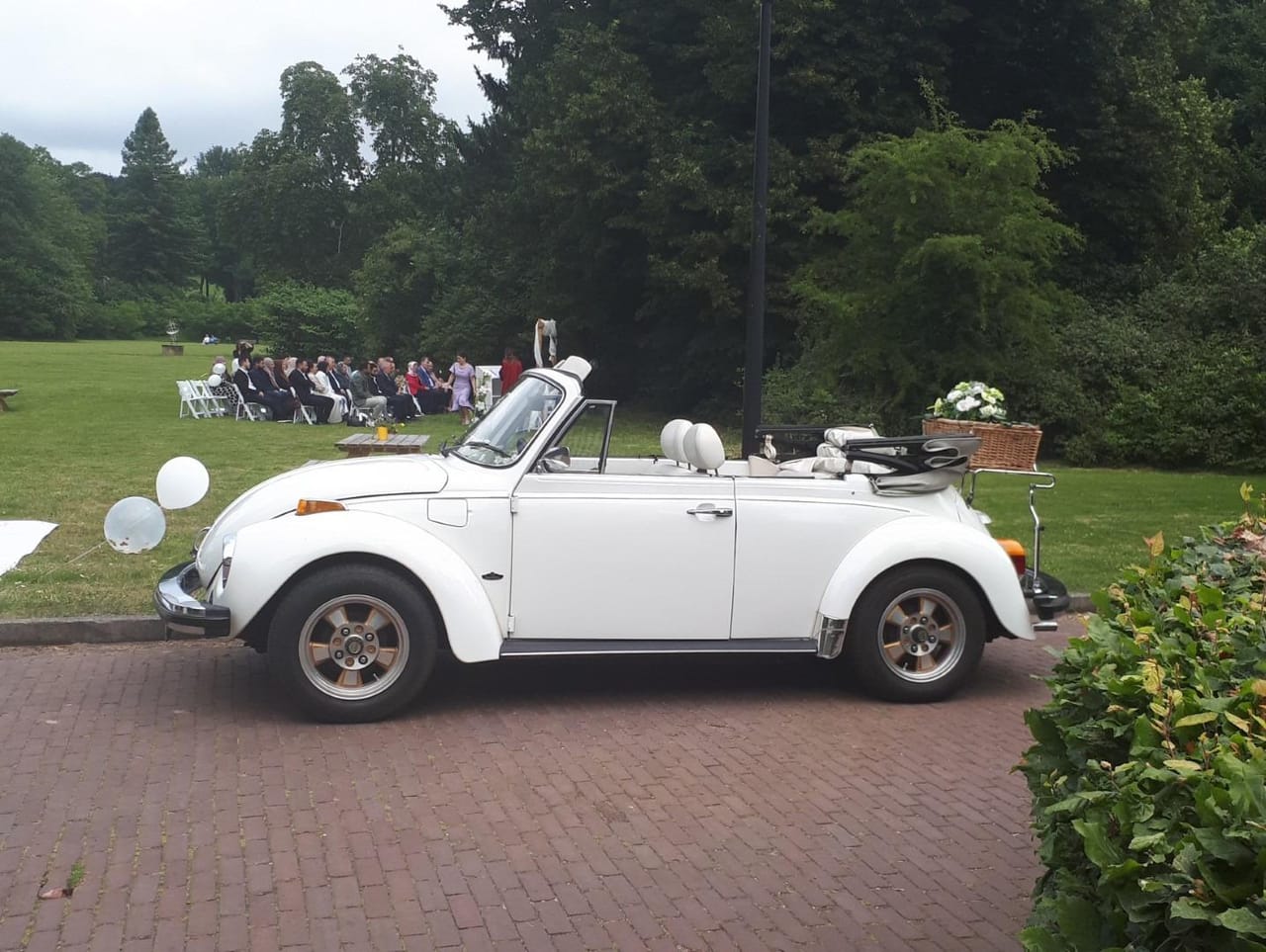 White Volkswagen Kever convertible with the roof down, balloons and a basket of flowers on the luggage rack, beside a lawn where guests are seated for an outdoor ceremony