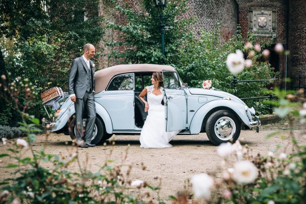 Light blue Volkswagen Kever convertible with a beige roof, a small bouquet of pink roses on the bonnet in front of the windscreen and a wicker basket with tulle at the back, the bride sitting in the open door and the groom standing beside it